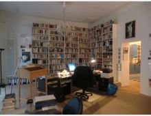 Arbeitszimmer mit großen, deckenhohen Bücherregalen, die drei Wände bedecken. Im Raum stehen ein Schreibtisch mit Computer und Stuhl sowie verschiedene Büromöbel; im Hintergrund ist ein Durchgang in einen angrenzenden Raum zu sehen.