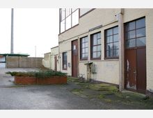 Gelände mit einem älteren Industriegebäude aus hellgelbem Backstein, großen Fenstern mit braunen Rahmen und einer braunen Tür. Im Vordergrund ein gepflasterter Hof mit einem bepflanzten gemauerten Hochbeet; im Hintergrund Holzzäune und ein niedriger Lagerraum.