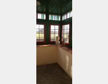 Ein kleiner Raum mit grünen Holzdecken und roten Fensterrahmen, durch die viel Licht einfällt. Die oberen Fensterbereiche haben dekorative Einsätze mit buntem Glas. Auf der Fensterbank stehen zwei Skulpturen, darunter eine Büste.