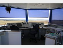Innenansicht eines Fluglotsen- oder Kontrollturms mit Blick auf eine Start- und Landebahn im Freien. Der Raum enthält Eingabegeräte wie Monitore, Schalter und Mikrofone sowie mehrere Bürostühle und Schreibtische.