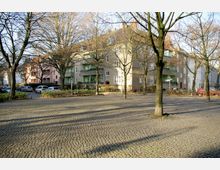 Mehrstöckiges Wohngebäude mit hellen, schlichten Fassaden und Balkonen, umgeben von Bäumen ohne Laub; im Vordergrund ein gepflasterter Platz mit vereinzelten parkenden Autos.