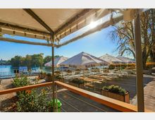 Außenbereich eines Biergartens mit Holzbänken und Tischen unter großen weißen Sonnenschirmen, mit Blick auf einen See und umliegende Bäume an einem sonnigen Tag. Im Vordergrund sind Pflanzen in Holzkästen sowie ein Geländer zu sehen.