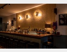 Eine stilvoll gestaltete Bar mit einer langen hölzernen Theke, auf der zahlreiche Flaschen, Gläser und Barutensilien stehen. Die Wand hinter der Theke ist mit gold-schimmernden Fliesen verkleidet und mit modernen Wandleuchten beleuchtet; daneben befinden sich Getränke- und Speisekarten.