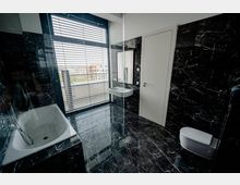 Modernes Badezimmer mit schwarzen Marmorböden und -wänden, einer Badewanne links, einer weißen Tür rechts und einem Fenster mit Jalousien, das Tageslicht hereinlässt. Ein Waschbecken mit Spiegel befindet sich neben der Tür, und rechts ist eine moderne Toilette sichtbar.