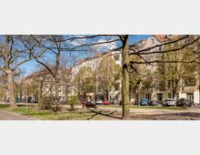 Straßenszene mit mehrstöckigen Wohnhäusern im Hintergrund, hellem Putz und roten Ziegeldächern, davor eine Allee mit Bäumen und einem kleinen Parkbereich mit Bänken. Geparkte Autos sind entlang der Straße sichtbar, unter blauem Himmel mit wenigen Wolken.