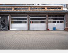Ehemaliges Backsteingebäude mit drei großen Garagentoren aus Glas und Metall, darüber schmale Fensterreihen. Links an der Fassade ist eine metallische Wandinstallation angebracht. Der Vordergrund besteht aus einem gepflasterten Hof.