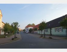 Straßenszene in einer Kleinstadt mit gepflastertem Gehweg, Bäumen und Häusern mit roten und schwarzen Ziegeldächern. Auf der rechten Seite ist ein mintgrünes Fachwerkhaus mit weißen Fensterrahmen und grünen Fensterläden zu sehen.