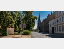 Straßenszene mit einer Baumallee auf der linken Seite und Wohngebäuden auf der rechten Seite, im Hintergrund ein Kirchturm mit spitzem Dach. Im Vordergrund stehen ein Stadtplan und ein Verkehrsschild am Rand eines Weges.