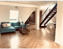 Ein modernes Wohnzimmer mit hellem Fischgrät-Holzboden, einer blauen Eckcouch, einem niedrigen Holztisch und einer Metalltreppe mit Holzstufen, die zu einem oberen Stockwerk führt. Im Hintergrund ist eine freiliegende Ziegelwand sichtbar.