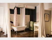 Ein Badezimmer mit freistehender, weißer Badewanne auf Holzfußboden, umgeben von Vorhängen. Im Hintergrund sind weiße, quadratische Fliesen zu sehen, ein Waschbecken mit Armaturen und ein grüner Paravent mit floralem Muster.