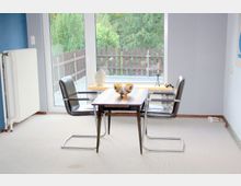 Ein kleiner Essbereich mit einem rechteckigen Holztisch und zwei schwarzen Stühlen aus Metall und Kunstleder. Im Hintergrund befindet sich eine große Fensterfront mit Blick auf eine Terrasse, einen Holzzaun und dichte Bäume.