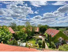 Hof mit mehreren roten Backsteingebäuden und roten Ziegeldächern, umgeben von Bäumen und gepflegten Gärten. Im Vordergrund ist ein Innenhof mit Gehwegen, Gartenmöbeln und einer Terrasse sichtbar, im Hintergrund erstreckt sich eine ländliche Landschaft unter einem sonnigen Himmel mit vereinzelten Wolken.