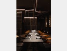 Modernes Restaurant mit dunklem, elegantem Interieur, langen Tischen, gedeckt mit Tellern und Gläsern, und Holzbänken mit gepolsterten Sitzen. Die Wände sind in dunklem Design gehalten, akzentuiert durch warme Beleuchtung und goldene Linien.
