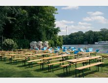 Eine Biergarten-Szene an einem Flussufer mit langen Holzbänken und Tischen, ergänzt durch blaue Liegestühle. Im Hintergrund sind Bäume, ein Schiff auf dem Fluss und ein Schornstein zu sehen.