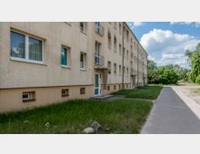 Mehrstöckiges Wohngebäude mit heller Fassade, kleinen Balkonen und einem Eingang mit einem Vordach. Vor dem Gebäude befindet sich eine Rasenfläche, angrenzend an einen Gehweg, umgeben von Bäumen im Hintergrund unter einem teils bewölkten Himmel.