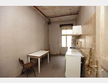 Schmale, einfache Küche mit abgewohntem Erscheinungsbild. Links ein Holztisch mit zwei Stühlen, rechts eine Küchenzeile mit Herd und Schränken, sowie beige Fliesen an der Wand; im Hintergrund ein großes einfaches Fenster.