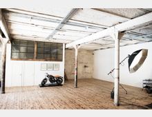 Industrieller Innenraum mit Holzfußboden, weißen Wänden und freiliegender Decke mit Metallträgern. Im Vordergrund eine Motorrad-Ausstellung, daneben ein professioneller Studioblitz und weitere Fotoausrüstung.