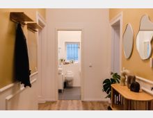 Flur mit hellgelben Wänden, Holzfußboden und einer Garderobe an der linken Wand. Rechts steht ein Holztisch mit Dekorationen und Spiegeln an der Wand. Im Hintergrund ist ein modernes Badezimmer mit weißer Einrichtung und einem Fenster sichtbar.