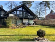 Ein modernes Einfamilienhaus mit schwarzem Holzrahmen und großen Glasfronten, flankiert von einem Garten mit Rasenfläche, Bäumen und einem kleinen Feuerkorb im Vordergrund. Im Hintergrund stehen weitere Häuser und Bäume, die Umgebung wirkt ländlich und ruhig.
