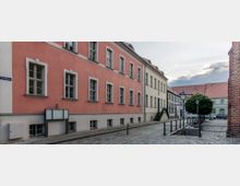 Straßenszene mit gepflastertem Gehweg, flankiert von historischen Gebäuden mit roten und cremefarbenen Fassaden. Im Vordergrund ein Baum, Laternen und eine kleine Brücke über einen Kanal.