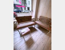 Zwei gepolsterte, verzierte Sitzbänke mit floralen Mustern und rosa Bezügen stehen auf einem hellen, abgenutzten Holzboden in einem Raum mit großen Fenstern. Draußen sind eine Straße mit parkenden Autos und die Fassade eines Gebäudes sichtbar.