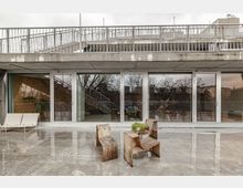 Moderne Terrasse mit Betonboden und Glasfassade, die den Blick nach draußen reflektiert. Vor der Fassade stehen schlichte Holzmöbel und Pflanzen, im Hintergrund ist ein Geländer mit Treppe zu sehen.