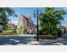 Straßenecke in einer Kleinstadt mit einem mehrstöckigen Gebäude mit Satteldach und Geschäften im Erdgeschoss, daneben bepflanztes Blumenbeet und große Bäume, im Hintergrund weitere Wohnhäuser. Ein nostalgischer Straßenlaternenmast steht im Vordergrund.