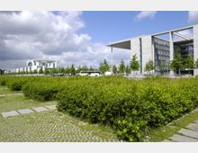 Moderne Regierungsgebäude in Berlin, umgeben von Grünflächen mit niedrigen Büschen und einem gepflasterten Steig. Im Hintergrund sind das Bundeskanzleramt und weitere moderne Architektur zu sehen, begleitet von einreihigen Bäumen und geparkten Fahrzeugen.