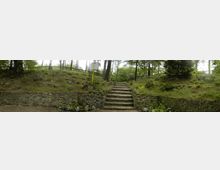 Steinerne Treppe, die einen bewaldeten Hügel hinaufführt, flankiert von niedrigen Natursteinmauern und umgeben von Gras, Farnen und Bäumen. Im Hintergrund ist ein leicht bewaldetes Gelände mit einem Zaun erkennbar.