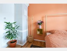 Schlafzimmer mit orangefarbener Wand, einem Bett mit farblich abgestimmter Bettwäsche, einer Topfpflanze neben einem weißen Kachelofen, einem gelben Regal mit Blumenvase und einem Holzständer. Der Boden besteht aus hellem Holzdielen.
