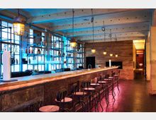 Moderne Bar in einem Industrie-Look mit einer langen Holztheke, hohen Barhockern und Regalen aus Metall und Glas an einer Backsteinwand. Hängende, industrielle Deckenlampen sorgen für warmes Licht, während blaue Akzentbeleuchtung die Wand hinter der Theke hervorhebt.