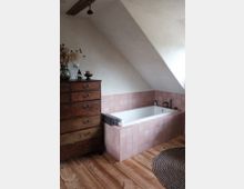 Dachgeschoss-Badezimmer mit einer weißen Badewanne, die von rosa Fliesen umgeben ist, Holzboden, einer antiken Holzkommode und einem runden, geflochtenen Teppich. Eine schräge Zimmerdecke mit sichtbarem Holzbalken ergänzt den Raum.
