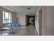 Flur eines modernen Gebäudes mit heller Wandfarbe und glänzendem Bodenbelag; links befinden sich große Fenster, unter denen blaue Wartestühle und Tische angeordnet sind, während rechts Bilder an der Wand hängen. Am Ende des Flurs ist eine Glastür mit einem beleuchteten Notausgangsschild erkennbar.