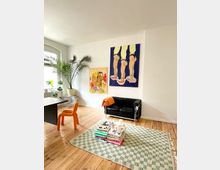 Ein heller Wohnraum mit Holzdielenboden, einer schwarzen Ledercouch, einem schwarzen Tisch, einem orangefarbenen Stuhl und grünen Zimmerpflanzen. An der weißen Wand hängen zwei farbenfrohe Kunstwerke, und auf einem gemusterten Teppich liegen gestapelte Bücher und Dekorationsgegenstände.