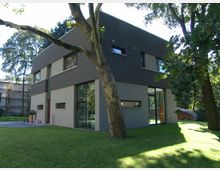 Modernes zweistöckiges Wohnhaus mit flachem Dach und klaren Linien, teilweise in dunkler und heller Fassade gestaltet, umgeben von einem gepflegten Rasen und Bäumen. Große Fenster und eine Glastür bieten Einblicke in den Innenraum, während im Hintergrund ein Baugerüst sichtbar ist.