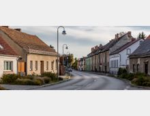 Straßenszene in einem Wohngebiet mit Reihenhäusern auf beiden Seiten, überwiegend in hellen Farben und mit Satteldächern. Die Straße ist schmal, leicht kurvig, mit Gehwegen und modernen Straßenlaternen versehen.