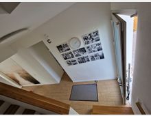 Treppenhaus einer modernen Wohnung mit hellen Wänden, Holzbodenstufen und Fliesenboden im Eingangsbereich. An der Wand befinden sich eine Uhr und eine Collage aus schwarz-weißen Fotos; eine geöffnete Haustür führt nach draußen.