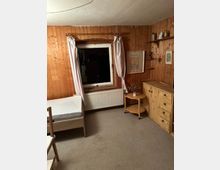 Schlafzimmer mit holzvertäfelten Wänden, einem Einzelbett mit Holzrahmen, einem Fenster mit weißen Heizkörpern darunter sowie hellrosa Vorhängen. An der rechten Wand befinden sich ein kleiner runder Beistelltisch, eine Kommode und wandmontierte Regale mit Dekorationsgegenständen. Der Boden ist mit beigem Teppich ausgelegt.