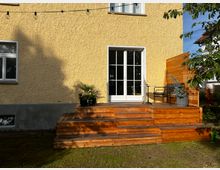 Gelbe Hausfassade mit einer weißen Terrassentür, die zu einer erhöhten Terrasse aus Holz führt. Die Terrasse hat Stufen, eine Sitzbank, einen kleinen Tisch und dekorative Pflanzen; im Vordergrund ist ein Gartenbereich mit Rasen zu sehen.