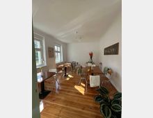 Heller Café- oder Bistro-Innenraum mit hohen weißen Wänden, großen Fenstern und poliertem Holzdielenboden. Mehrere kleine Tische mit Metall- und Holzstühlen stehen verteilt; an der Wand hängen Bilder, und auf einem Tisch steht eine Vase mit roten Blumen.