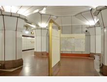 U-Bahn-Station mit weißen Wandverkleidungen und roten Akzenten, mehreren breiten Säulen und einer goldenen Infotafel in der Mitte. An der Wand hängt ein Stadtplan, und im Hintergrund ist der Schriftzug „Altstadt Spandau“ zu sehen.