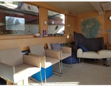 Ein gemütlicher Innenraum mit hellem Holz verkleideten Wänden, mehreren Polsterstühlen, einem weißen Sessel und Regalen mit Büchern. Große Fenster geben den Blick auf den rostigen Rumpf eines Außenschiffes frei, während eine abstrakte Kunstgrafik eine der Wände dekoriert.