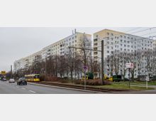 Wohnsiedlung mit mehreren weiß-grauen Plattenbauten, flankiert von einer mehrspurigen Straße und Straßenbahngleisen. Im Vordergrund stehen Bäume ohne Laub, ein gelber Straßenbahnwagen und parkende Fahrzeuge; Schilder und Verkehrsschilder sind ebenfalls sichtbar.