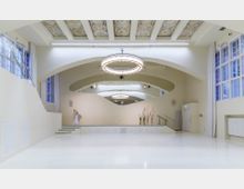 Innenraum eines modernen Veranstaltungsraums mit heller, geschwungener Raumstruktur und großer, weißer, glänzender Bodenfläche. An der Decke befinden sich dekorative Elemente und ein großer, runder Kronleuchter; an den Seiten sind hohe Fenster und Vorhänge zu sehen.