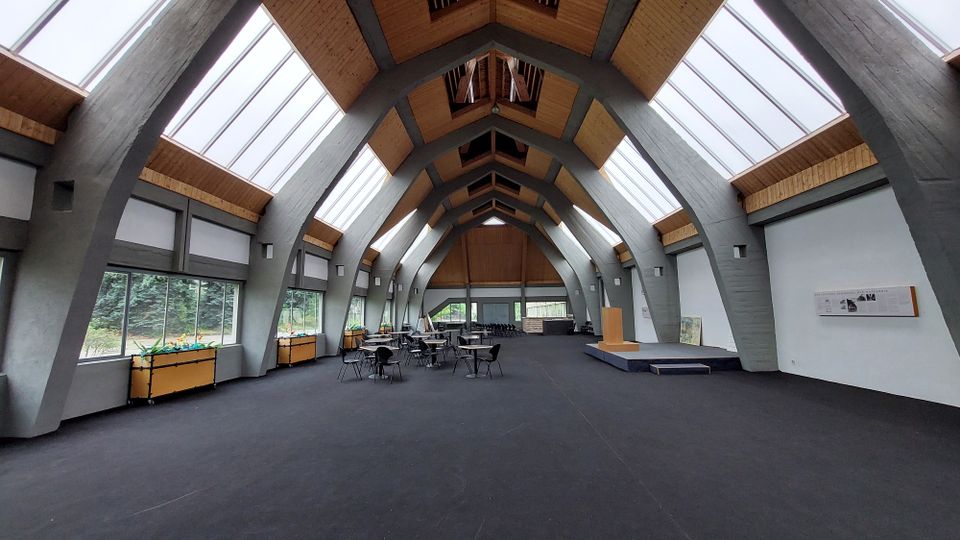 Großer, moderner Innenraum mit einer gewölbten Decke aus Holz und Betonträgern, seitlich große Fenster mit Blick ins Grüne. Der Raum ist mit vereinzelten Tischen, Stühlen und einem kleinen Podium ausgestattet, möglicherweise genutzt für Veranstaltungen.
