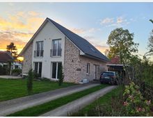 Ein modernes Einfamilienhaus mit weißer und rötlicher Klinkerfassade, Satteldach und bodentiefen Fenstern. Im Vordergrund ist ein gepflasterter Weg, der zu einer Garage mit geparktem Auto führt, umgeben von Rasen und Bäumen, während der Himmel im Hintergrund durch den Sonnenuntergang farbig erleuchtet ist.