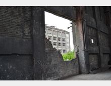 Teilansicht eines verfallenen Industriegebäudes mit grauen, beschädigten Ziegelwänden und einem großen Fenster ohne Glas. Durch die Öffnung ist ein weiteres verlassenes Gebäude mit zerbrochenen Fenstern sowie Vegetation im Hintergrund sichtbar.