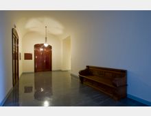 Langer, schlichter Flur mit glänzendem grauen Boden, cremefarbenen Wänden und Holzakzenten. Im Hintergrund befinden sich zwei hölzerne Türen mit der Beschriftung „Saal N“ und „Sitzung“, daneben eine hölzerne Sitzbank an der rechten Wand.