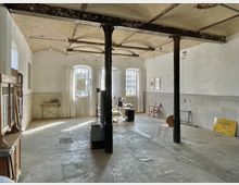 Industrieähnlicher Raum mit hohen Decken, sichtbaren Metallträgern und großen Fenstern mit weißen Vorhängen, die Tageslicht einlassen. Im Raum befinden sich einzelne Möbel wie ein Holzschrank, Stühle und technische Geräte, der Boden ist aus Beton.