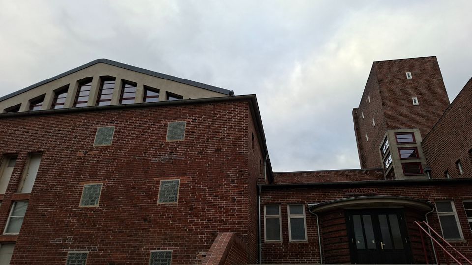 Backstein-Gebäude mit schlichtem, funktionalem Design, bestehend aus mehreren Segmenten mit kleinen Fenstern. Ein gewölbter Eingangsbogen sowie ein hoher Turm im Hintergrund dominieren die Architektur.